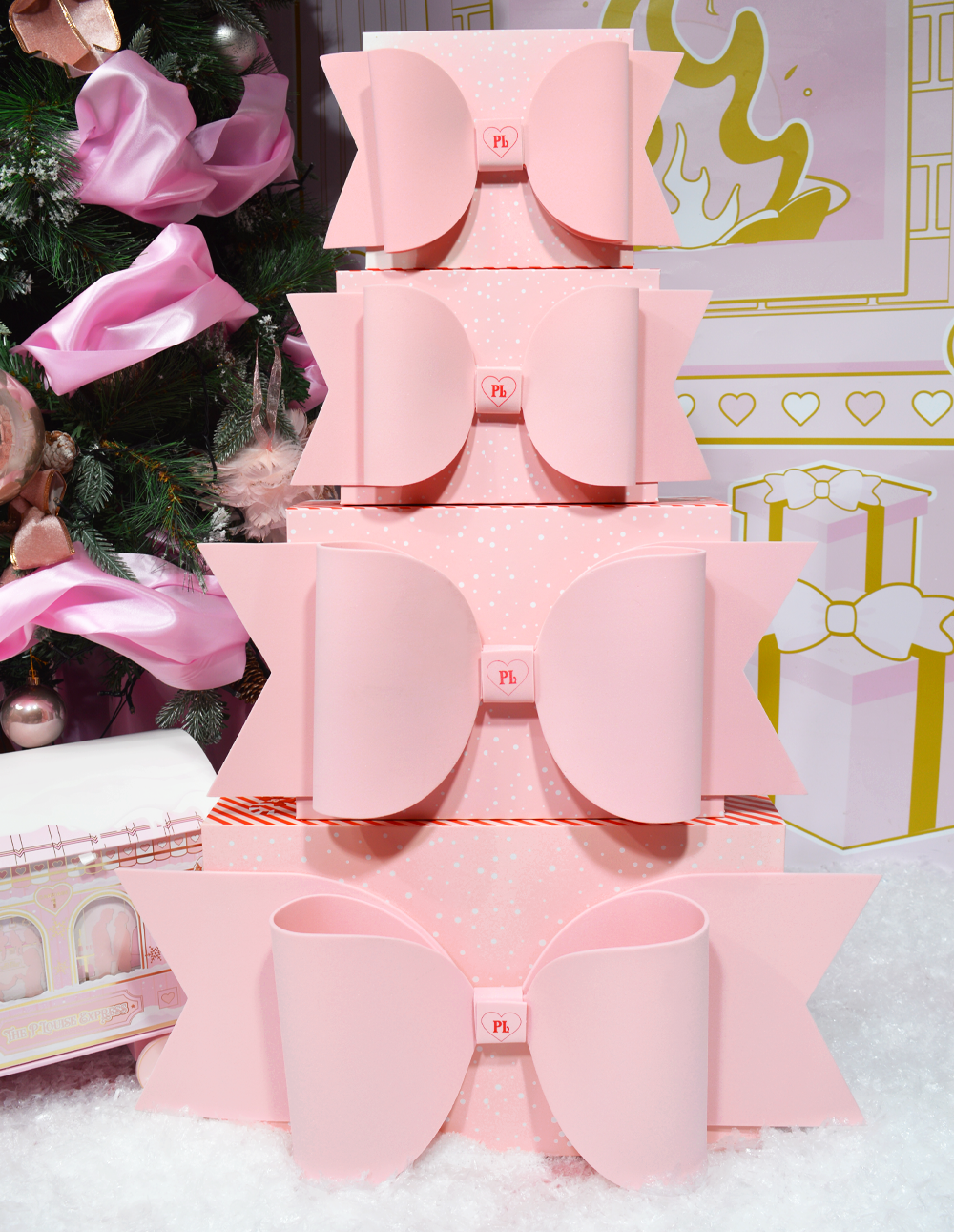 กองกล่องห่อของขวัญ P.Louise สีชมพูพร้อมโบว์ตั้งโชว์โดยมีฉากหลังเป็นธีมคริสต์มาสพร้อมของตกแต่ง