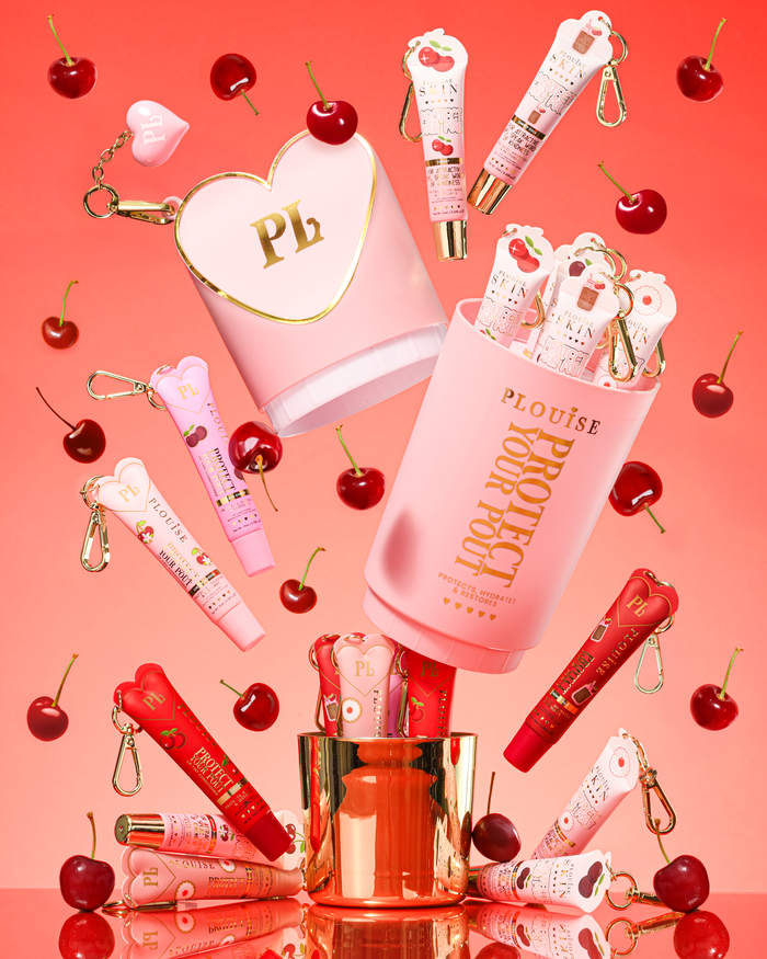 Pink XL Protect Your Pout Gift Set ที่เปิดล้อมรอบด้วยเชอร์รี่และครีมนวดริมฝีปากสีชมพูและสีแดงบนพื้นหลังสีชมพู