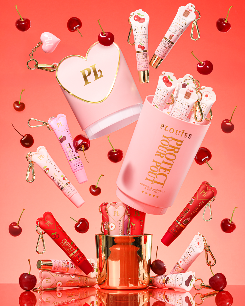 Pink XL Protect Your Pout Gift Set ที่เปิดล้อมรอบด้วยเชอร์รี่และครีมนวดริมฝีปากสีชมพูและสีแดงบนพื้นหลังสีชมพู