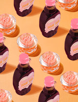 ภาชนะใสของ P.Louise Dose of Dreams Vitamin C Capsule Cream โดยมีแคปซูลสีส้มอยู่ข้างใน ล้อมรอบด้วยขวดน้ำเชื่อมสีม่วงและสีชมพูบนพื้นสีส้ม
