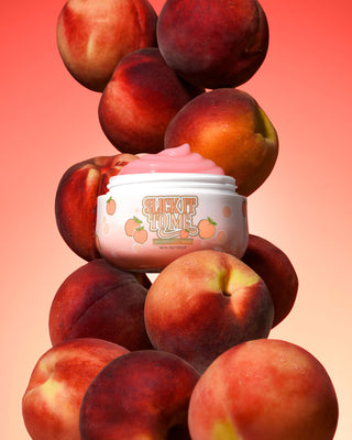 Jar of P. Louise Slick It To Me Peach Melting Body Balm พร้อมครีมสีชมพูล้อมรอบด้วยลูกพีชบนพื้นหลังแบบไล่ระดับ