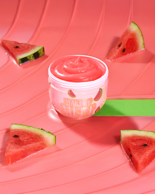 Open tub of P. Louise Watermelon Make It Melt Makeup Cleansing Balm surrounded by watermelons on a red background