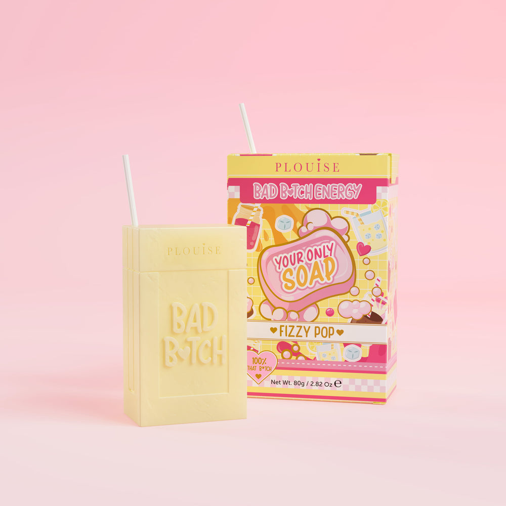 สบู่ก้อนสีเหลืองที่มีคำว่า 'Bad Bitch' สลักอยู่บนนั้น พร้อมด้วยกล่องบรรจุน้ำผลไม้หลากสีสันที่มีป้ายกำกับ 'Fizzy Pop' อยู่บนพื้นหลังสีชมพู
