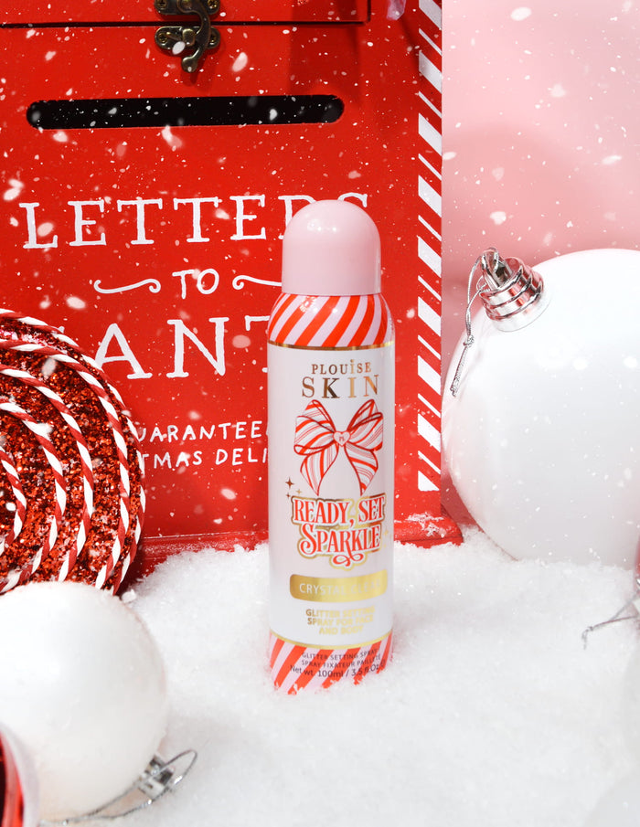ขวด P. Louise Ready Set Sparkle Christmas Glitter Setting Spray ล้อมรอบด้วยของประดับตกแต่งตามเทศกาล เช่น หิมะ ของประดับตกแต่ง และกล่องจดหมายคริสต์มาส