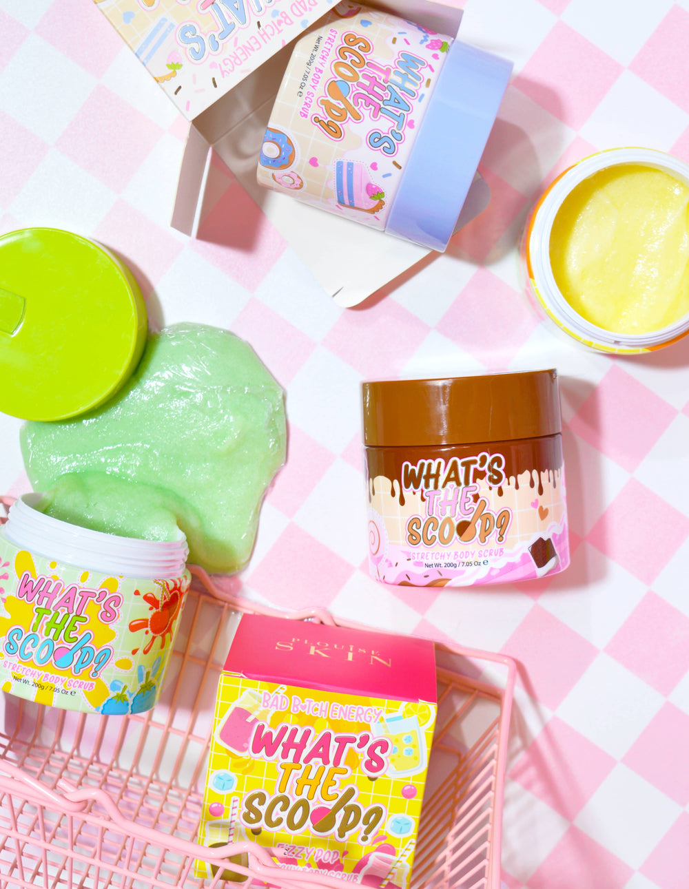 กล่องใส่สครับขัดผิวหลากสีสันหลากสีสันพร้อมชื่อผลิตภัณฑ์ 'What's the Scoop?' ปรากฏให้เห็นเด่นชัด บรรจุภัณฑ์ถูกจัดเรียงบนพื้นผิวที่มีพื้นหลังลายตารางหมากรุกสีชมพู และมีกราฟิกและข้อความที่สนุกสนานและมีสีสันที่เกี่ยวข้องกับผลิตภัณฑ์