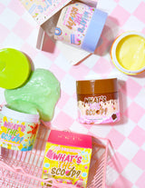 กล่องใส่สครับขัดผิวหลากสีสันหลากสีสันพร้อมชื่อผลิตภัณฑ์ 'What's the Scoop?' ปรากฏให้เห็นเด่นชัด บรรจุภัณฑ์ถูกจัดเรียงบนพื้นผิวที่มีพื้นหลังลายตารางหมากรุกสีชมพู และมีกราฟิกและข้อความที่สนุกสนานและมีสีสันที่เกี่ยวข้องกับผลิตภัณฑ์